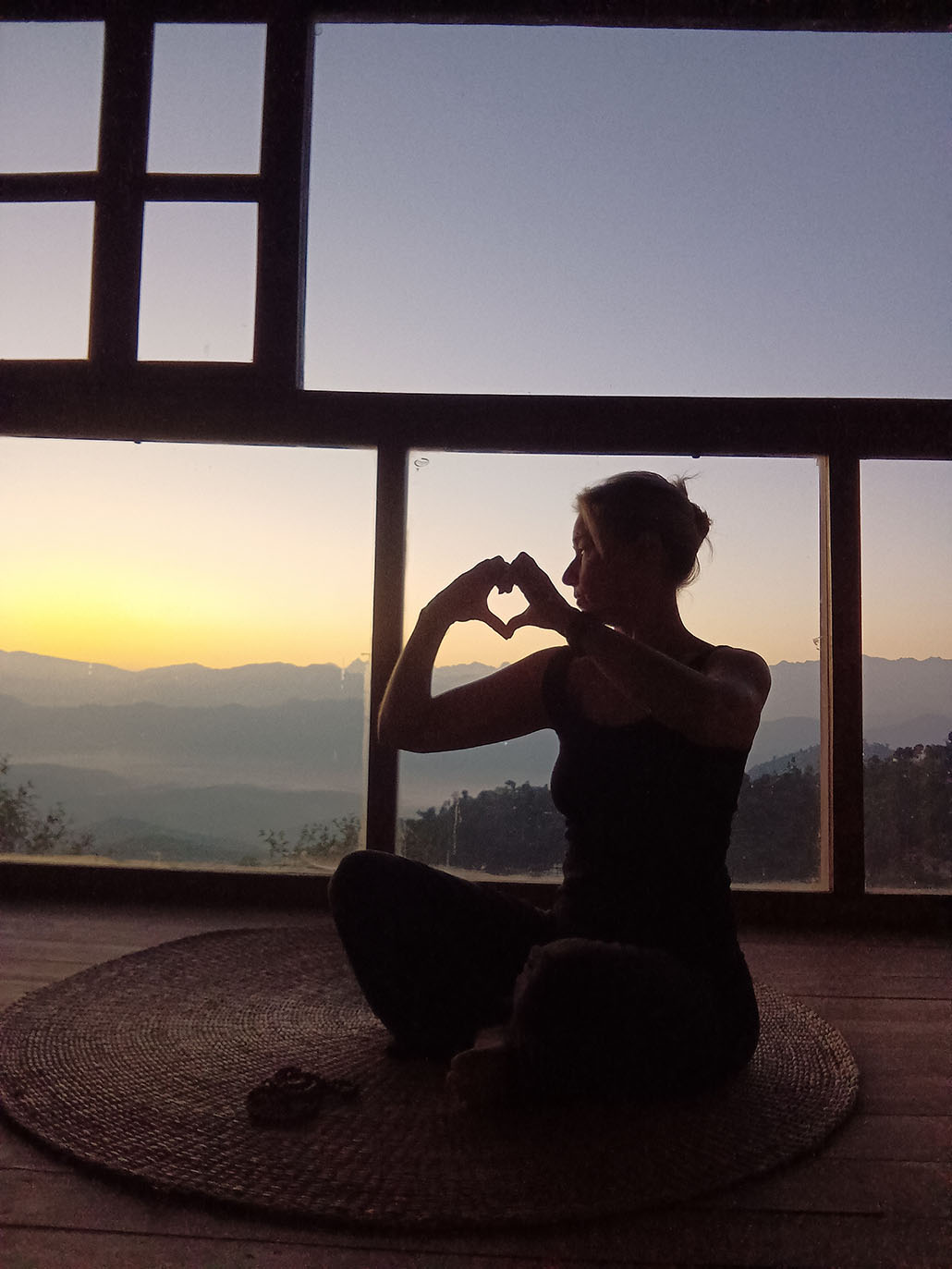 Istuva nainen tekee sydänkuvion käsillään ja katsoo ikkunasta auringonnousua