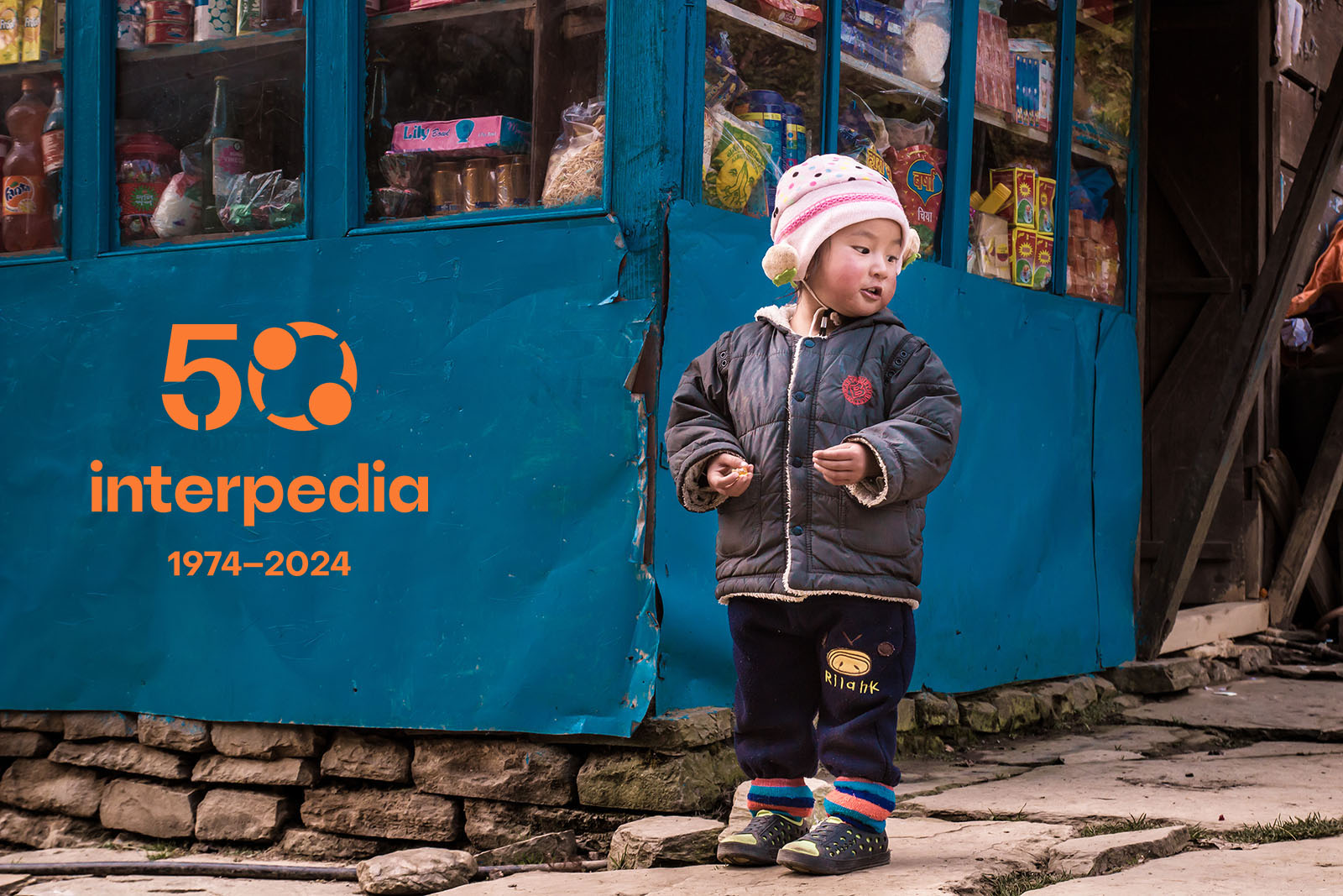 Nepalilainen lapsi seisoo kadulla kaupan edessä. Kuvassa on myös Interpedian 50-vuotisjuhlan logo